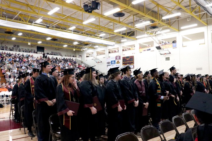 
              A large group of students in graduation caps and gowns stand in rows, facing forward during a commencement ceremony held in a gymnasium. The graduates wear maroon and gold stoles, some holding diplomas. The gym is filled with attendees seated in the bleachers.